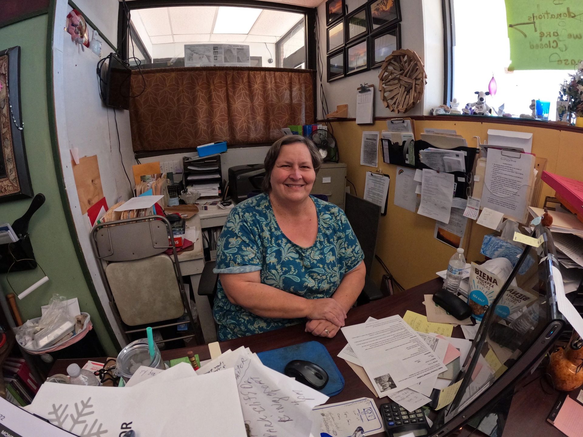 A volunteer smiling behind a desk covered in paperwork inside the pantry office